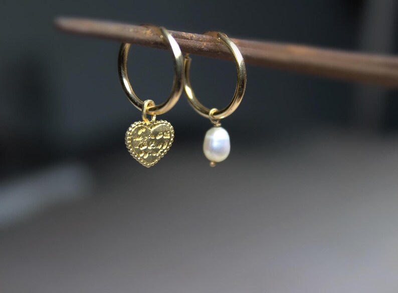 May include: A pair of gold hoop earrings with a charm dangling from each. One charm is a heart-shaped pendant with the words "Love you to the moon and back" inscribed on it. The other charm is a white pearl.