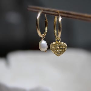 May include: Two gold hoop earrings with charms. One earring has a small white pearl charm, and the other has a gold heart charm with the words "Peace Love Joy" inscribed on it.