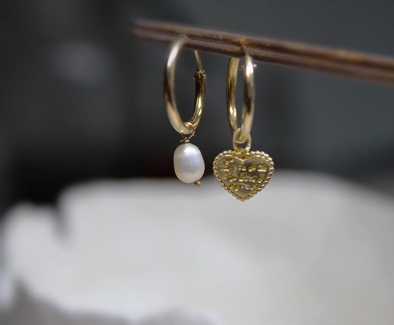 May include: Two gold hoop earrings with charms. One earring has a small white pearl charm, and the other has a gold heart charm with the words "Peace Love Joy" inscribed on it.