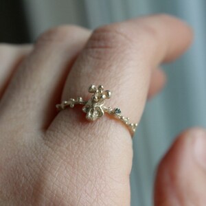 May include: A gold ring with a delicate, organic design featuring a small cluster of gold beads and a single black gemstone. The ring is worn on a person's finger.