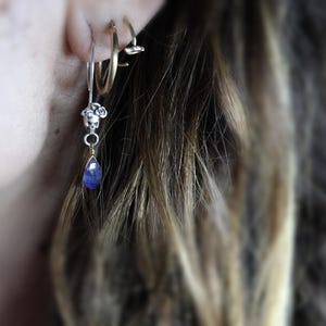 May include: A silver hoop earring with a small silver charm dangling from it. The charm features a cluster of small white beads and a teardrop-shaped blue gemstone.