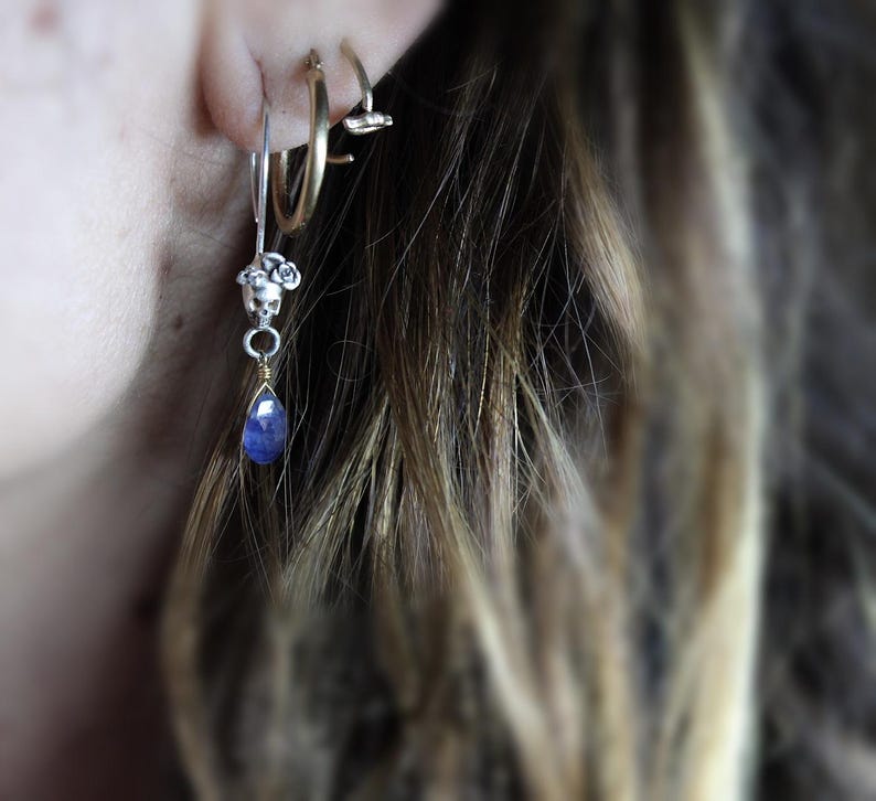 May include: A silver hoop earring with a small silver charm dangling from it. The charm features a cluster of small white beads and a teardrop-shaped blue gemstone.