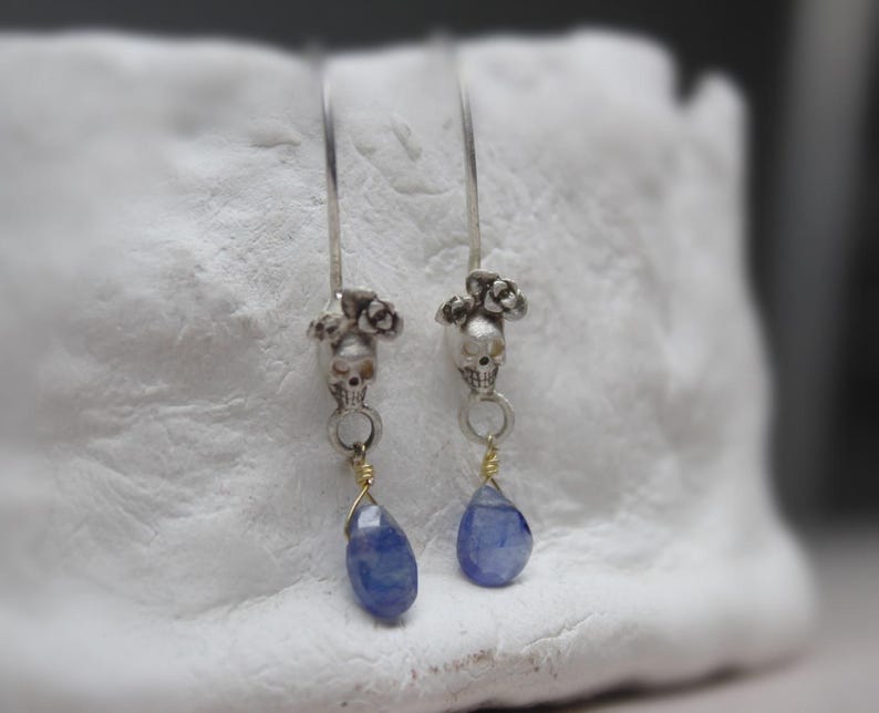 May include: Silver skull earrings with blue teardrop stones hanging from the bottom. The skulls are adorned with small silver flowers.