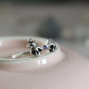 May include: A silver ring with two skull-shaped charms and a small blue gemstone in between.