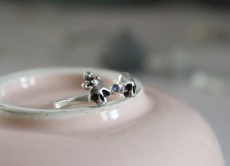 May include: A silver ring with two skull-shaped charms and a small blue gemstone in between.