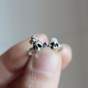 May include: A silver ring with two small skull charms and a blue gemstone in the center.