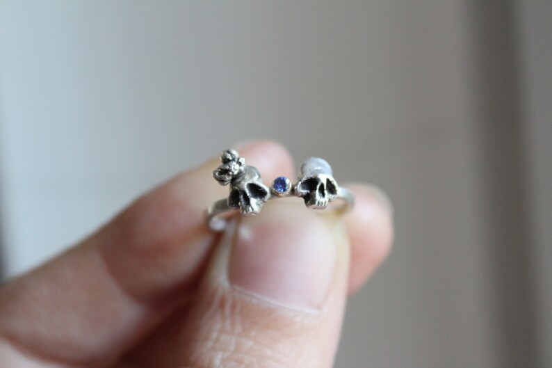 May include: A silver ring with two small skull charms and a blue gemstone in the center.