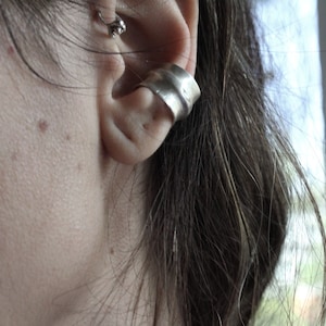 Puede incluir: Un brazalete de plata para la oreja envuelve el cartílago de la oreja. El brazalete tiene un diseño orgánico texturizado. También se ve un pequeño pendiente de plata en la parte superior de la oreja. La tez de la persona es clara y su cabello es oscuro.