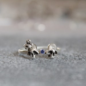 May include: A silver ring with two skull-shaped designs and a small blue gemstone in the center.