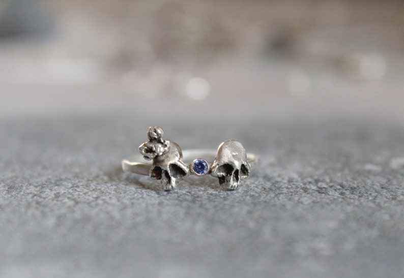 May include: A silver ring with two skull-shaped designs and a small blue gemstone in the center.