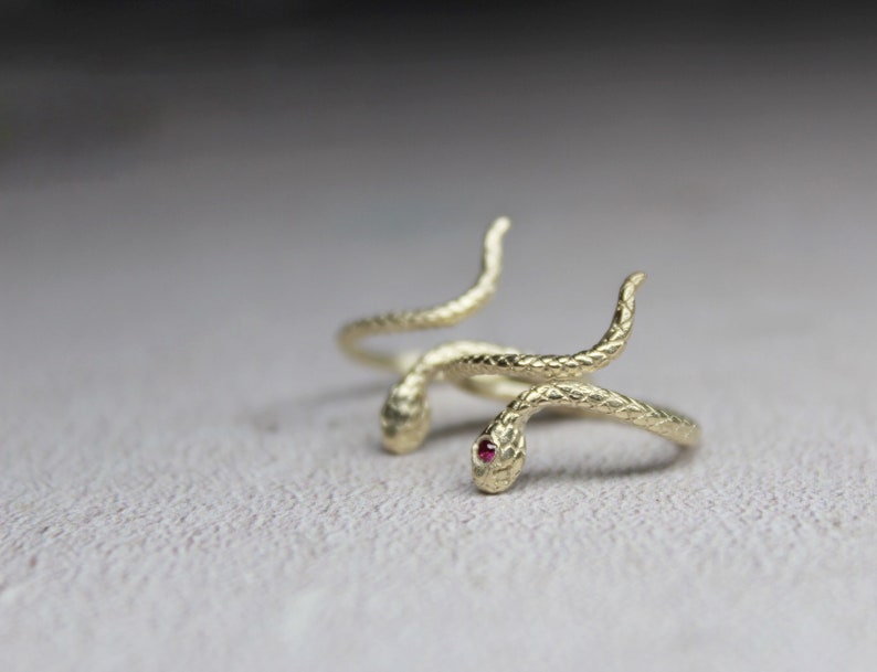 May include: A gold snake ring with a red gemstone eye. The snake is coiled around itself, with the tail wrapping around the finger.