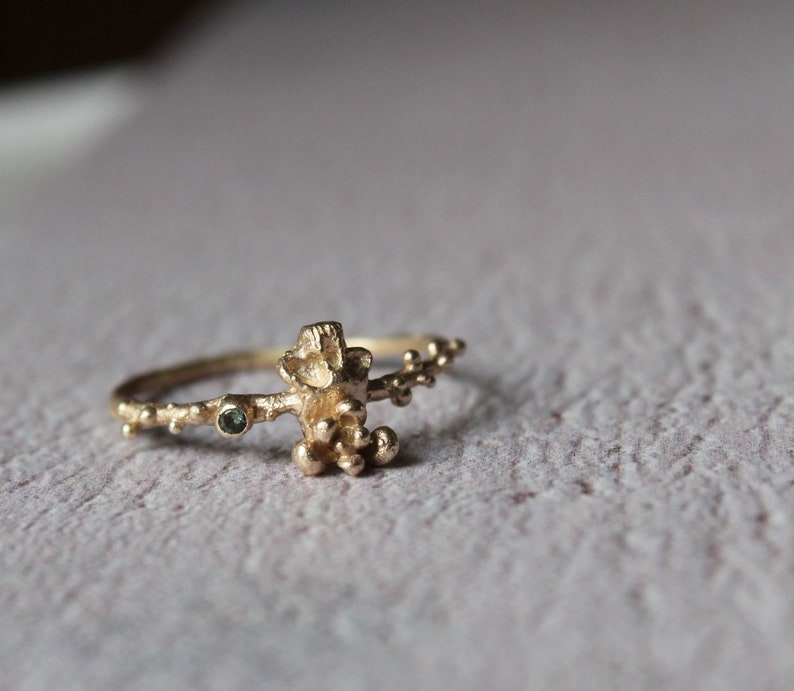 May include: A gold ring with a delicate, organic design featuring a cluster of small, round stones and a single, larger green stone. The ring is made of a textured metal and has a unique, natural look.