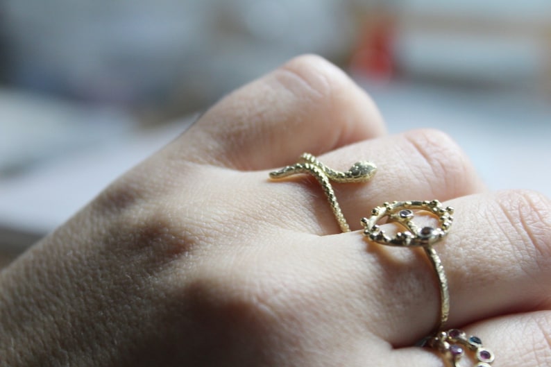 May include: A hand wearing three gold rings. The top ring is a snake design, the middle ring is a crown with small gemstones, and the bottom ring is a thin band with small gemstones.