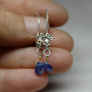 May include: Silver skull earrings with blue teardrop-shaped stones hanging from them. The skulls have floral details.