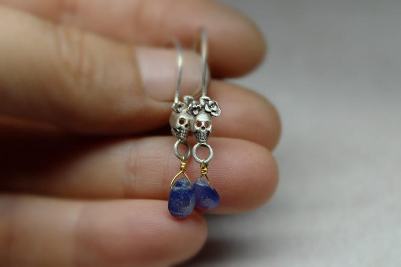 May include: Silver skull earrings with blue teardrop-shaped stones hanging from them. The skulls have floral details.