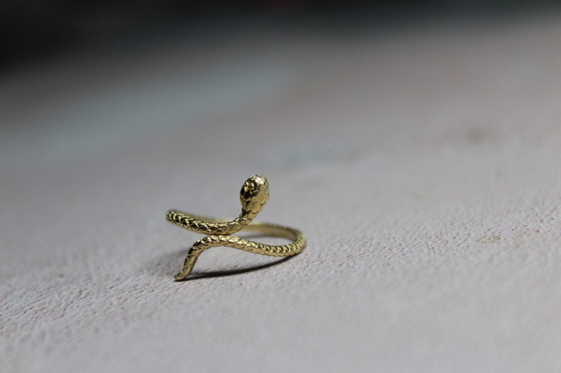 May include: A gold snake ring with a detailed, textured design. The snake is curled around the ring band, with its head raised and facing forward.
