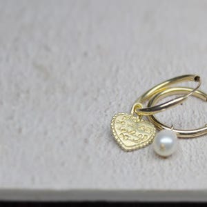 May include: Gold hoop earrings with a heart charm and a pearl dangle. The heart charm has an inscription on it.