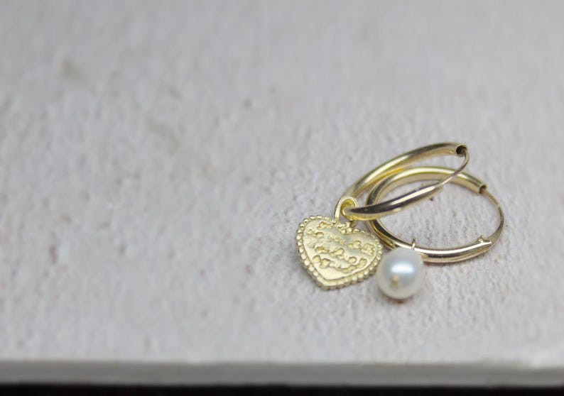 May include: Gold hoop earrings with a heart charm and a pearl dangle. The heart charm has an inscription on it.