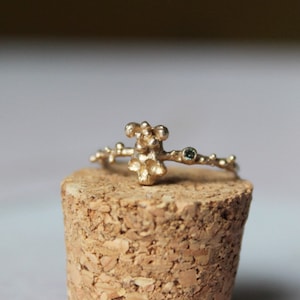 May include: A gold ring with a textured, organic design featuring a cluster of small spheres. The ring is adorned with a single, small, green gemstone.