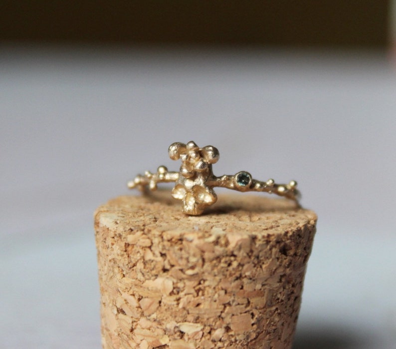 May include: A gold ring with a textured, organic design featuring a cluster of small spheres. The ring is adorned with a single, small, green gemstone.