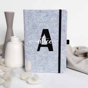 Könnte beinhalten: Ein graues Filzjournal mit einem schwarzen Gummibandverschluss. Das Cover zeigt einen großen schwarzen Buchstaben "A" mit dem Namen "alice" in Schreibschrift. Das Journal ist rechteckig und minimalistisch gestaltet.