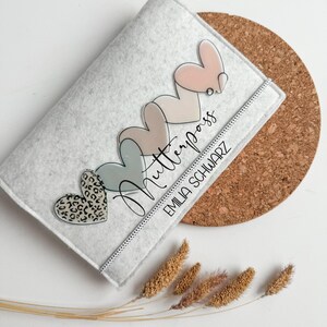Könnte beinhalten: Eine graue Filztasche mit Reißverschluss. Die Tasche hat ein Design aus fünf Herzen in verschiedenen Rosatönen und Blau, mit einem Leopardenmuster-Herz unten. Der Text "Mutterpass" ist in Schreibschrift geschrieben, und der Name "EMILIA SCHWARZ" ist darunter gedruckt.