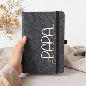Könnte beinhalten: Ein dunkelgraues Filzjournal mit dem Wort "PAPA" in Weiß und einem Namen in goldener Schrift. Das Journal hat einen schwarzen Gummibandverschluss. Das Journal wird von einer Hand gehalten.