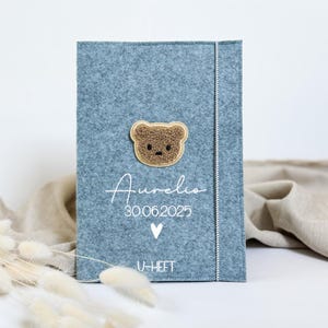 Könnte beinhalten: Eine blaue Babybuchhülle aus Filz mit einem braunen Teddybär-Applikation. Der Einband trägt den Namen "Aurelio", das Datum "30.06.2025", ein Herzsymbol und den Text "U-HEFT" in Weiß. Ein weißer, gestickter Rand verläuft entlang der rechten Seite.