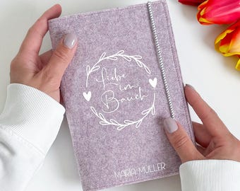 May include: A lavender felt journal cover with the words "Liebe im Bauch" in a white floral design. The journal cover has a white elastic band closure and the name "Maria Müller" printed at the bottom.