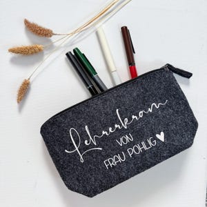 Könnte beinhalten: Ein dunkelgraues Filz-Federmäppchen mit schwarzem Reißverschluss, gefüllt mit Stiften in verschiedenen Farben. Der Text "Lehrerkram von Frau Pohlig" ist in weißer Schrift geschrieben. Getrocknete Weizenähren sind im Hintergrund.