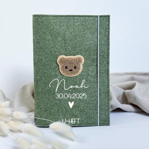 Könnte beinhalten: Eine grüne Filzhülle mit einem braunen Teddybär-Applikation. Der Name "Noah" und das Datum "30.04.2025" sind in weißer Schrift zusammen mit einem Herzsymbol aufgedruckt. Der Text "U-HEFT" ist unten aufgedruckt.