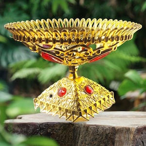 May include: An ornate gold-coloured pedestal bowl with red accents. The bowl features intricate filigree designs and is adorned with red jewels. The bowl is elevated on a square base with more red jewels and decorative edges. The bowl is likely used for decorative purposes.