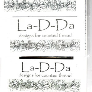 May include: A black and white image of a logo for a counted thread design company called "La-D-Da". The logo features a stylized floral border.