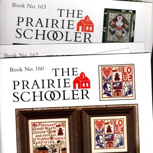 May include: Three covers of The Prairie Schooler cross-stitch pattern books. The covers are white with red and black text. The text on the covers reads "The Prairie Schooler" and "Book No. 163", "Book No. 162", and "Book No. 160". Each cover features a red schoolhouse illustration.