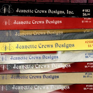 May include: A stack of colorful boxes with the text "Jeanette Crews Designs" in a decorative font. The boxes are in various colors, including black, red, yellow, and blue. Each box has a unique product number and price.
