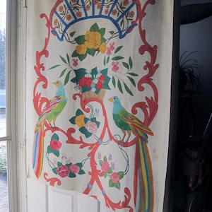 May include: A white fabric wall hanging with a colorful floral and bird design. The design features two birds with long, rainbow-colored tails, flowers, and leaves. The fabric is decorated with a red, blue, and white pattern. The bottom right corner of the fabric has the word "Fides" written in red.