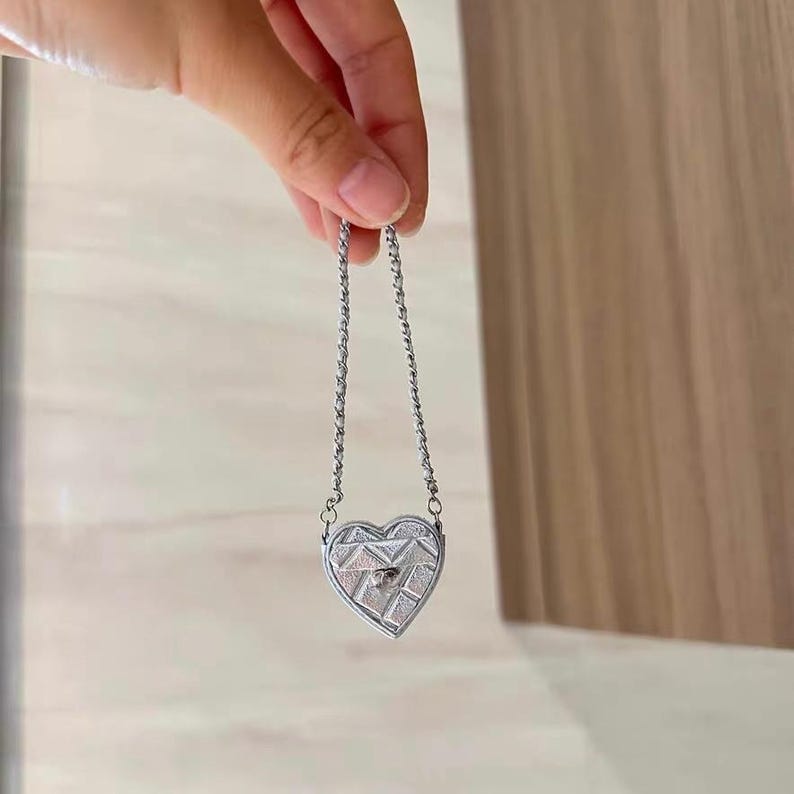 May include: A silver heart-shaped pendant necklace with a geometric design. The pendant is suspended from a delicate chain. The heart has a textured surface and a small, round gem in the center.
