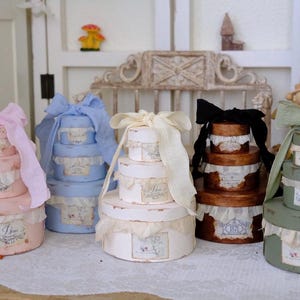 May include: A collection of decorative round boxes in various pastel colors, including pink, blue, white, brown, and green. Each box is stacked and adorned with a matching ribbon and lace trim. The boxes have a vintage aesthetic.