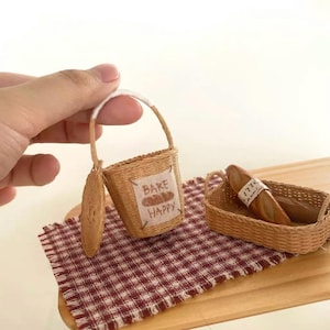 May include: A miniature wicker basket with a white handle and a small woven tag that reads "Bake Happy" with a bread graphic. A small woven plate and a basket with two miniature baguettes are also present. The items are on a red and white checkered cloth.