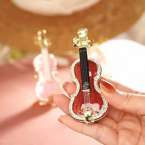 Könnte beinhalten: Eine Miniatur-Geigenfigur mit rotem Korpus, goldenen Akzenten und einem Rand aus klaren Kristallen. Die Saiten sind sichtbar, und eine weitere rosa Geige ist im Hintergrund leicht unscharf.