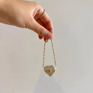 May include: A small, heart-shaped purse with a gold chain strap. The purse is silver with a quilted pattern and a gold clasp. The chain is made of small, gold-colored links. The purse is held by a hand with a silver ring.