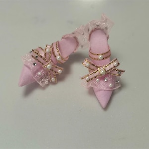 Op de afbeelding: Een paar miniatuur roze hoge hakken met gouden randen, strikken en parelaccenten. De hakken zijn gemaakt van stof en hebben een puntige neus. De strikken zijn versierd met parels en gouden details, wat een vleugje elegantie toevoegt.