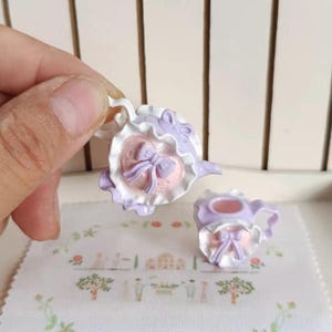 Puede incluir: Un juego de tetera y crema miniatura en colores pastel. La tetera y la crema son de color lavanda y blanco con detalles en rosa y lazos. El juego está diseñado con un estilo con volantes y adornado, perfecto para una casa de muñecas o exhibición decorativa.