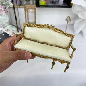 May include: A miniature gold-coloured sofa with a cream-coloured patterned seat and back. The sofa has ornate detailing and curved armrests. The legs are slender and gold. A small, decorative piece for a dollhouse or display.