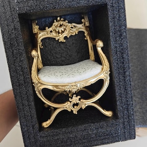 May include: A miniature gold-colored chair with ornate detailing, including curved legs and a decorative back. The seat is upholstered in a white fabric with a subtle pattern. The chair is presented in a protective foam insert.