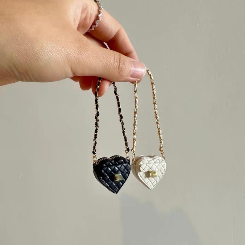 May include: Two heart-shaped mini purses, one black and one white, are suspended by chain straps. The purses have a quilted pattern and gold-colored hardware. The black purse has a black chain strap, while the white purse has a gold chain strap.