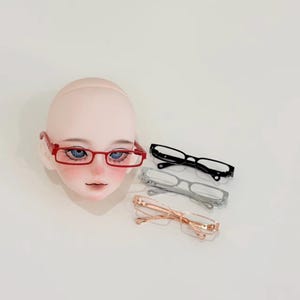 May include: A doll head with blue eyes wearing red rectangular glasses. Three additional pairs of glasses in black, silver, and rose gold are displayed. The glasses have a minimalist design.