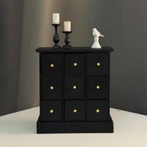 Może przedstawiać: Czarna miniaturowa komoda z dziewięcioma kwadratowymi szufladami, każda ze złotym guzikiem. Dwa czarne świeczniki z białymi świecami i biała figurka ptaka na górze. Komoda stoi na białej powierzchni.