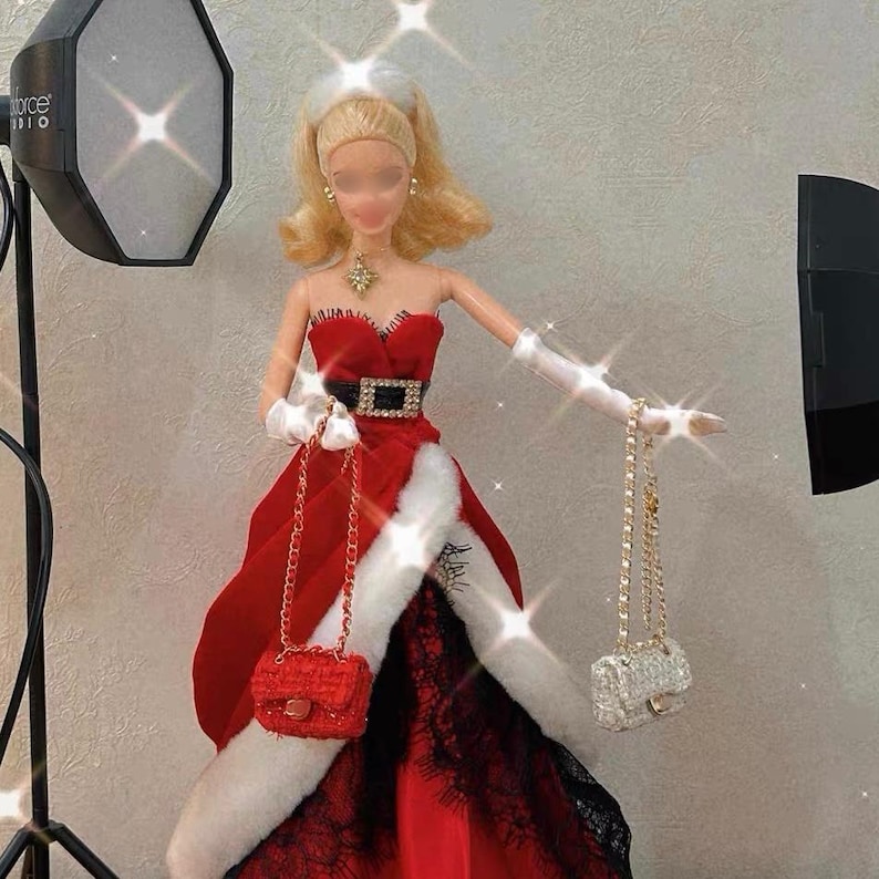 Puede incluir: Una mu&ntilde;eca con un vestido rojo sin tirantes con cintur&oacute;n negro y ribete de piel sint&eacute;tica blanca. La mu&ntilde;eca sostiene dos peque&ntilde;os bolsos, uno rojo y otro plateado, con correas de cadena dorada. La mu&ntilde;eca tiene el pelo rubio y lleva guantes blancos.