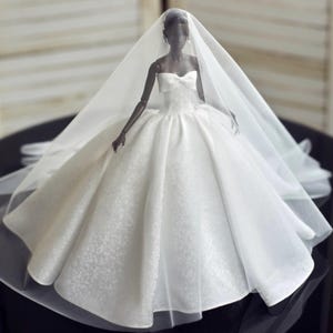 May include: A doll in a white strapless wedding gown with a bow detail and a full skirt. The doll is wearing a sheer veil. The doll's skin is dark, and it is posed in a formal setting. The gown's fabric has a subtle floral pattern.
